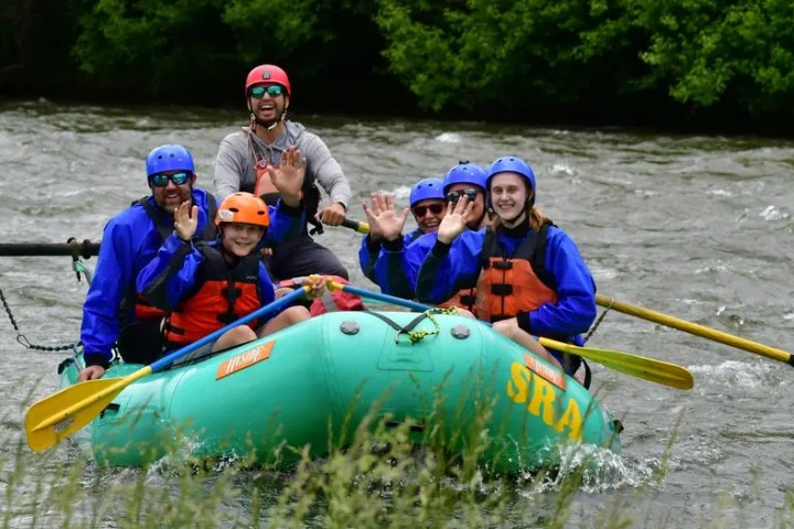 Group of people whitewater rafting, smiling and waving, wearing helmets and life jackets.