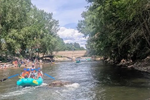 People rafting on a river with trees and a bridge in the background.