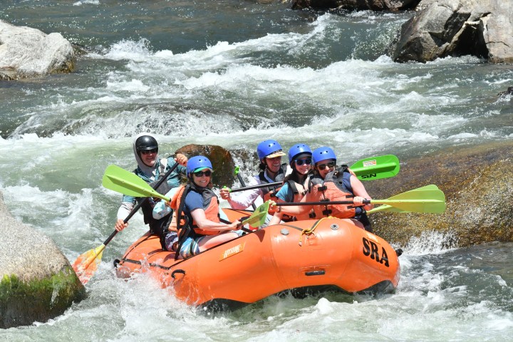 Group rafting on a rocky river in an orange inflatable raft, wearing helmets and life jackets.