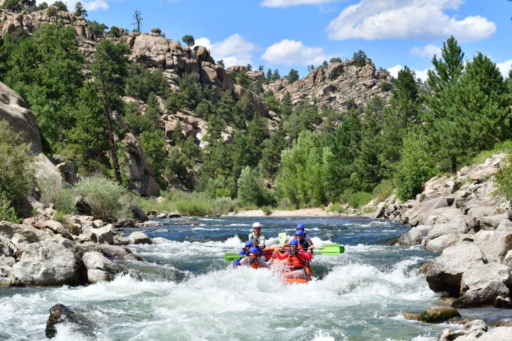 Group rafting through rapids on a rocky river with trees and clear sky.