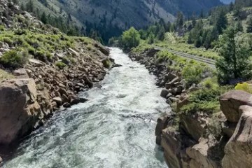 Rushing river flowing through rocky, forested canyon under clear sky.