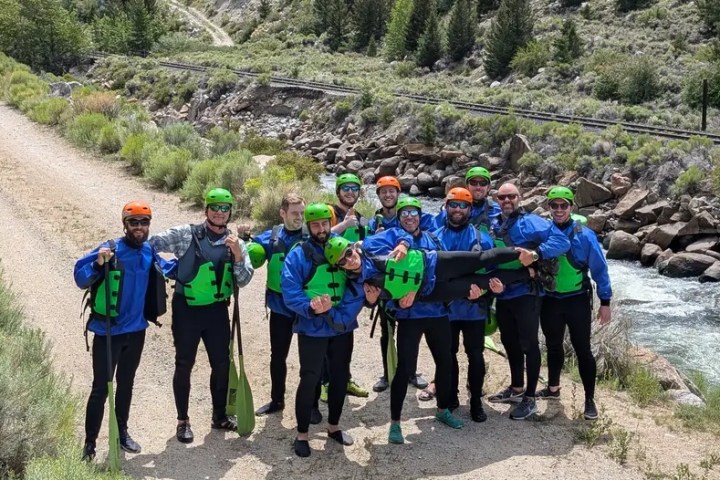 Group in blue jackets and helmets posing by a river, holding a person horizontally.