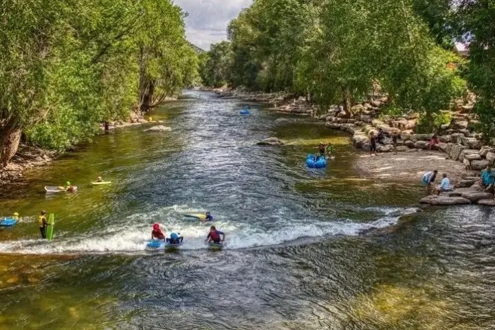 People enjoying tubing and relaxing by a tree-lined river on a sunny day.
