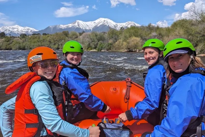 Four people in helmets and life jackets rafting on a river with snow-capped mountains in the background.