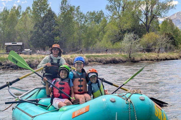Group of four people in safety gear rafting on a river with trees in the background.