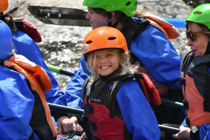 Group of people in helmets and vests rafting on a river, smiling and paddling.