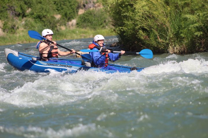 Two people kayaking in a blue tandem kayak on a whitewater river.
