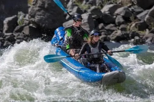 Two people kayaking in a blue inflatable kayak on a river with rapids.