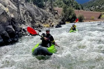 Two people kayaking down a rocky river with green inflatable kayaks and red paddles.