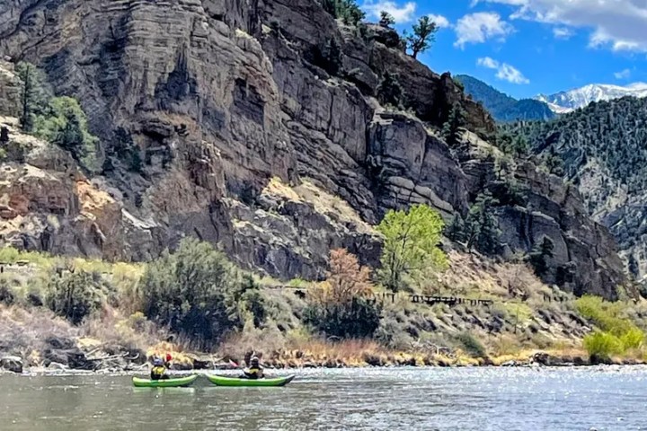 Two kayakers on river with rocky cliffs and trees under a partly cloudy sky.
