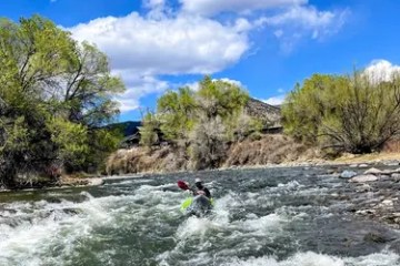 Person kayaking in a river with trees and blue sky in the background.